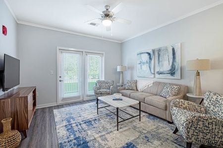 2 Bedroom Apartments in Santa Rosa Beach, FL - The Southern - Living Room with Wood-Style Flooring, Area Rug, Ceiling Fan, Couch, Armchairs, Coffee Table, End Tables with Lamps, Mounted TV, Entertainment Center, Floor-to-Ceiling Window with Blinds, and Glass Door with Access to Balcony.